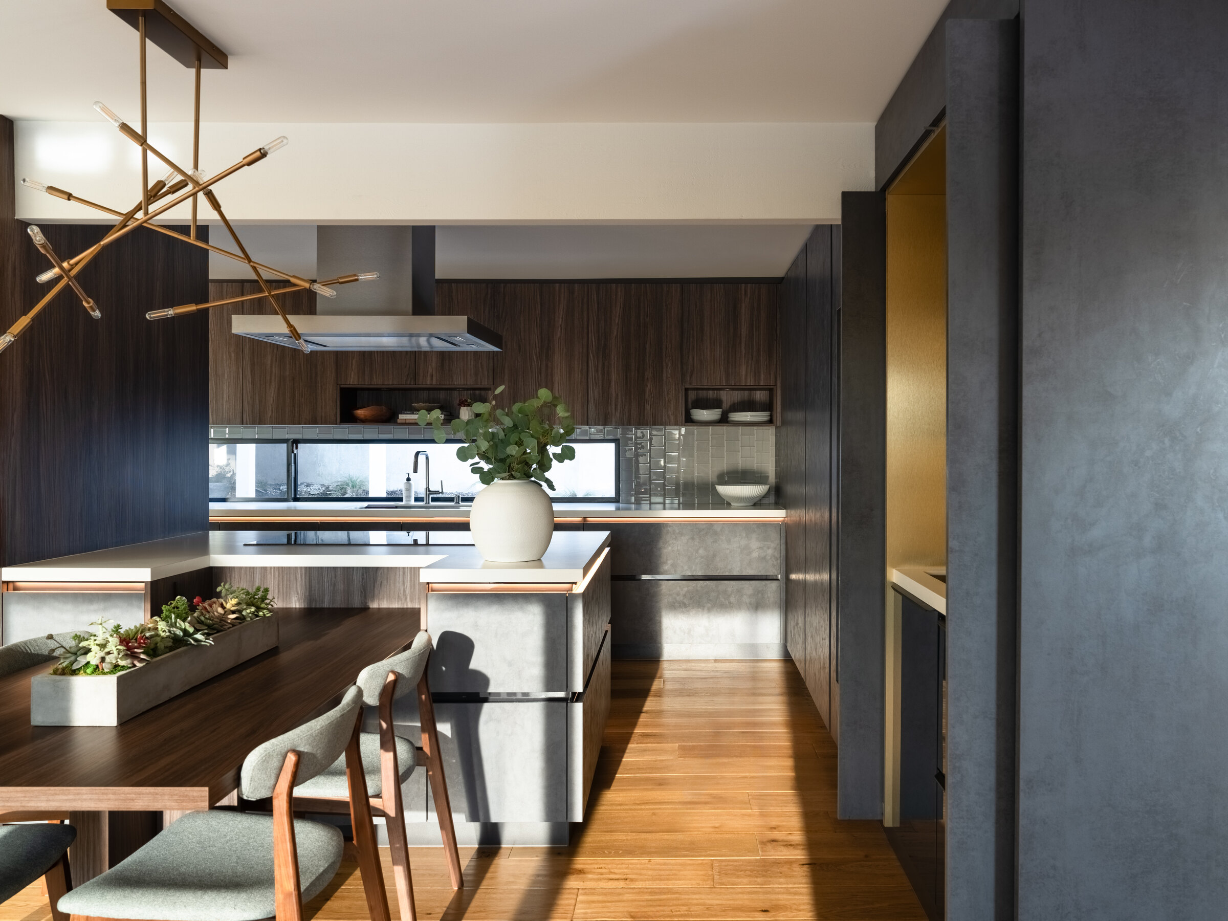Completed contemporary kitchen with dark wood cabinetry, central island, dining table, and sculptural brass lighting